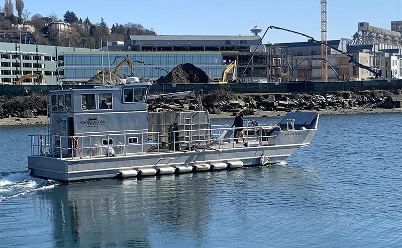 A metal workboat travels across calm water near a shoreline with construction equipment, buildings, and piles of material in the background.