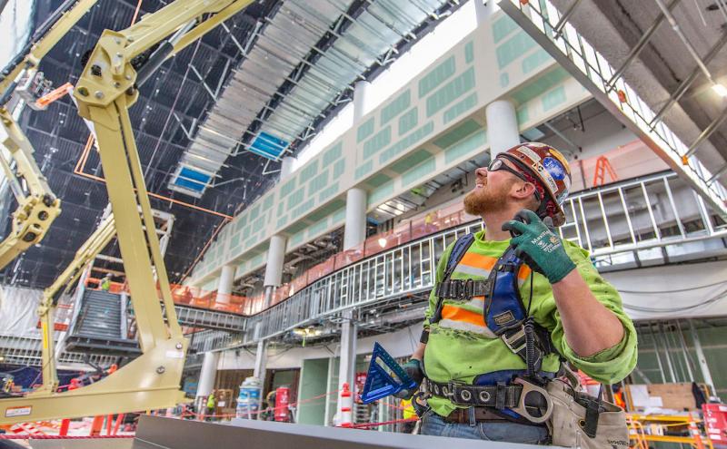 A construction worker in safety gear stands inside a large industrial building under construction with overhead machinery and metal framing.