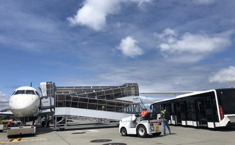 An aircraft parked on the airport ramp with a mobile boarding ramp attached, an airport bus positioned nearby, and ground crew working beside a small tow vehicle under a partly cloudy sky