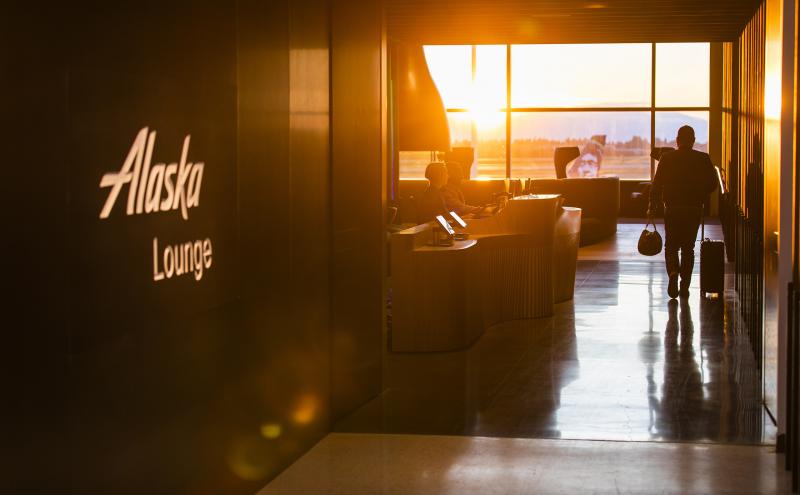 Sunlit entrance to the Alaska Lounge with a person walking down the corridor toward large windows at sunset.