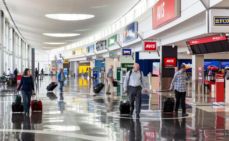 Inside an airport rental car area, several people walk with rolling luggage across a wide, glossy floor. Rental car counters with signs for Avis, Budget, National, Hertz, Alamo, and other companies line the right side of the space, while bright overhead lighting and large windows illuminate the scene.