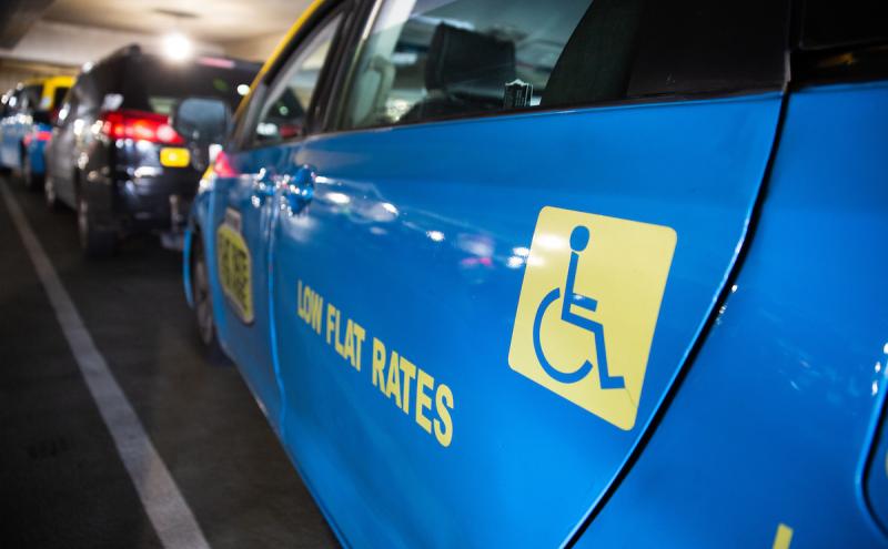 A row of taxis parked in a covered area, with a bright blue taxi in the foreground displaying a wheelchair accessibility symbol and the words “LOW FLAT RATES” on its side.
