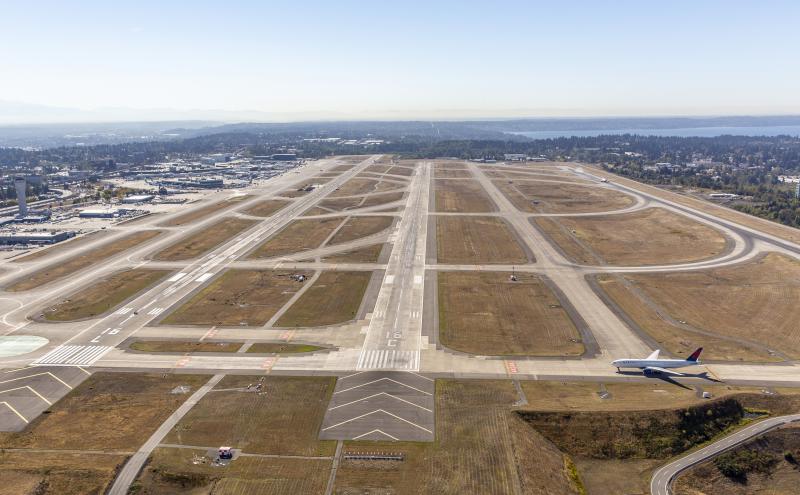 Aerial view of multiple Sea Airport runways with a commercial airplane taxiing and airport buildings and landscape visible in the background.