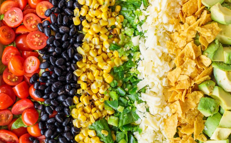 A close-up view of neatly arranged salad ingredients in horizontal rows, including sliced cherry tomatoes, black beans, corn kernels, chopped green bell peppers, shredded cheese, crushed tortilla chips, and diced avocado.