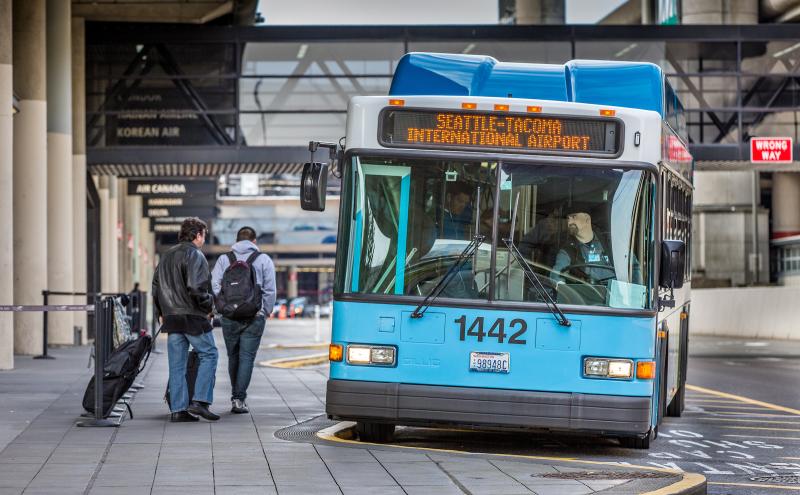 An airport shuttle bus marked ‘1442’ with a sign reading ‘Seattle–Tacoma International Airport’ is stopped at a curbside pickup area. Two people with luggage and backpacks walk toward the bus along a covered sidewalk lined with airline signage.
