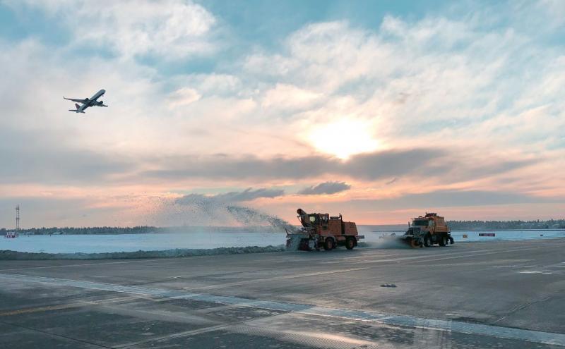 Snowplows at SEA plow the runways while a plane takes off with a sunset.
