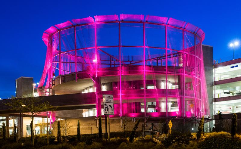 Circular airport parking structure illuminated in bright pink lighting at night, with the mesh-like exterior glowing pink and a deep blue sky in the background. Landscaping, roadway signs, and a nearby parking garage are visible in the foreground.