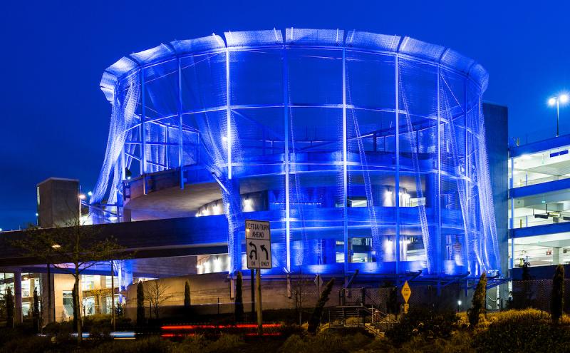 A circular multi-story structure wrapped in hanging strands of wire mesh draped from the roof to the ground, lit from within at night. 