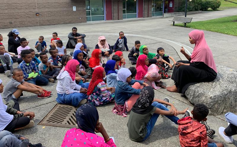A group of children seated outdoors in a semicircle on a paved area, listening to an adult who is sitting on a large rock and speaking to them. A building with windows and doors is in the background, along with a bench and grassy area.