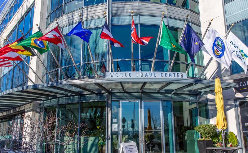 Exterior view of the World Trade Center Seattle building, featuring a curved glass façade and a row of international and organizational flags displayed above the main entrance. The entrance canopy, reflective windows, and nearby café umbrellas and signage are visible.