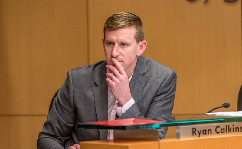 Commissioner Calkins seated at a meeting desk in a formal setting, wearing a suit and resting one hand near the chin. A nameplate reading “Ryan Calkins” is visible on the desk, along with papers and a glass panel, against a wood‑paneled background.