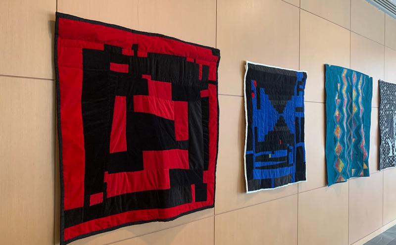 Various colored, patterned quilts hang on the wall.