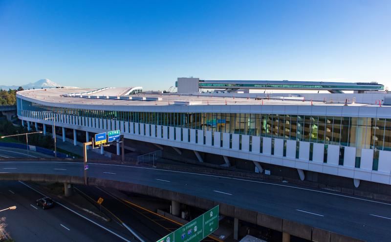 "Wide-angle view of a large modern airport terminal building with a curved roof and rows of tall glass windows. Multiple elevated roadways and highway signs run in front of the structure. Construction cones and equipment are visible on the rooftop. A clear blue sky fills the background, with a snow-covered mountain visible in the distance.