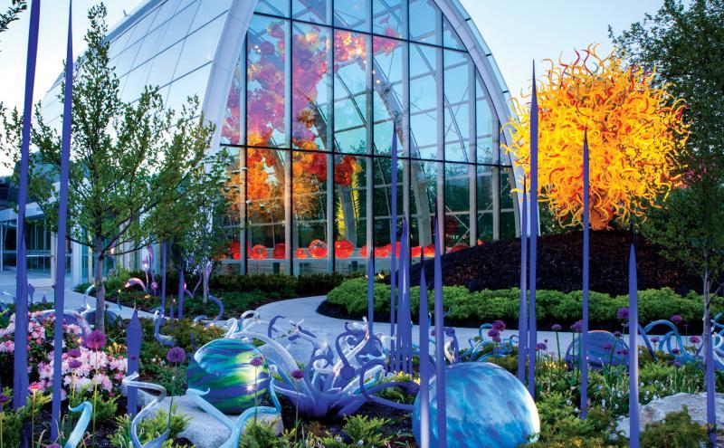 Outdoor garden featuring colorful glass sculptures among flowers and greenery. In the background, a large glass conservatory displays vibrant orange and red glass artwork inside, reflected on its curved windows. In the foreground, tall slender purple glass forms and rounded blue‑green glass shapes are arranged within landscaped plant beds.