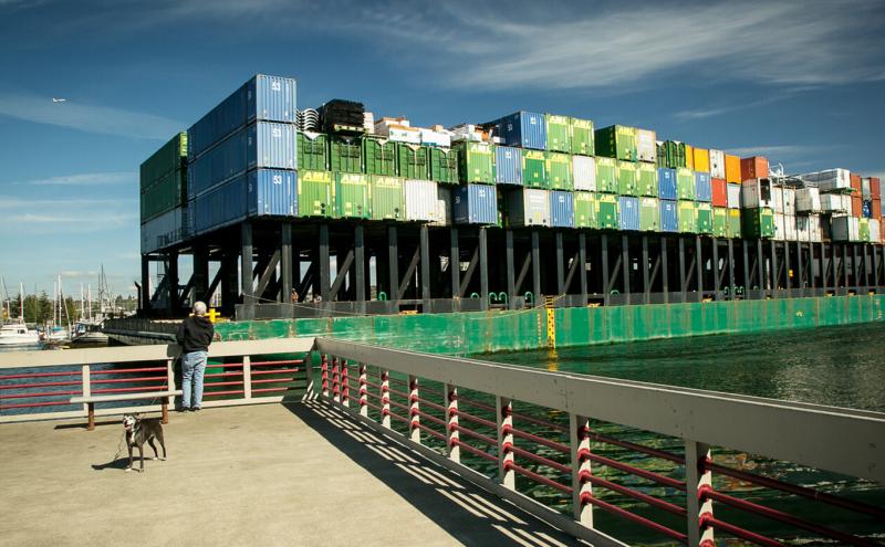A man and his dog look toward the Duwamish River where a barge with containers glides by. August, 2018, Seattle.