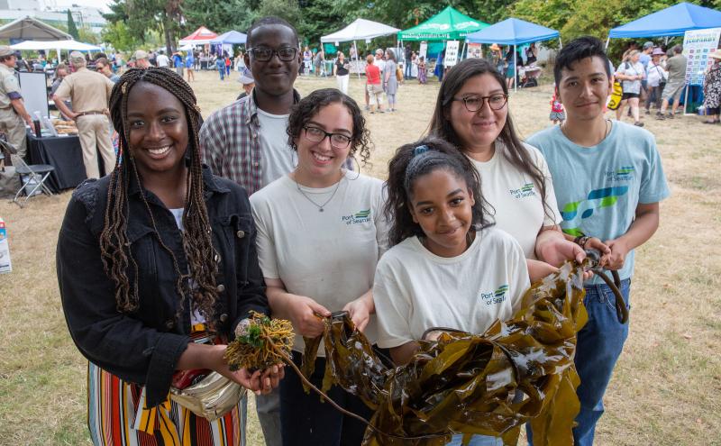 A group of people stands outdoors at a community event, holding large pieces of seaweed. Behind them are booths, tents, and other attendees in an open grassy area.