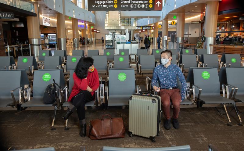 Two travelers seated in an airport waiting area with spaced‑apart chairs and directional signs visible overhead.