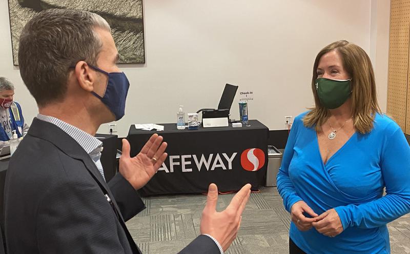 Two people stand in conversation in a room with a table covered by a black Safeway‑branded tablecloth. The table displays various items including bottles, papers, and a small sign that reads “Check‑in.” A large piece of artwork hangs on the wall, and the room has neutral-colored flooring and walls.