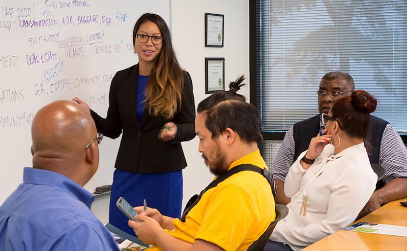 Mixed group of employees in a meeting with woman at a whiteboard leading the meeting.