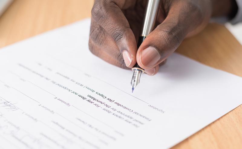 Hand signing a document on a light wood desk.