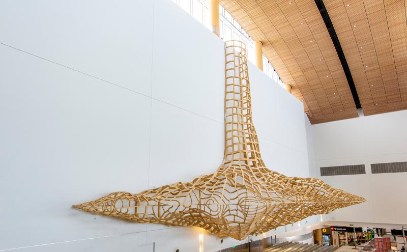 Tall, lattice-style wooden sculpture extending from the ceiling down a white wall inside an airport concourse.