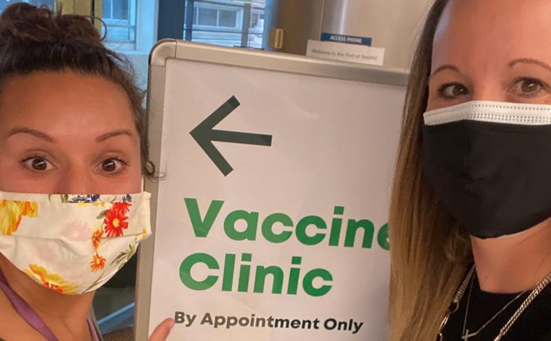 Two masked staff members along side sign pointing to a COVID-19 Vaccine Clinic at SEA Airport.