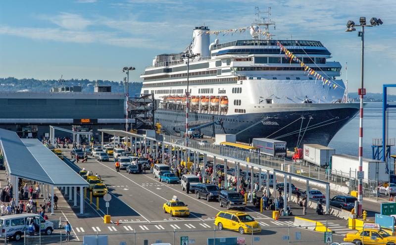 Large cruise ship docked at a busy terminal with long lines of taxis, cars, and passengers moving through loading and drop‑off areas under covered walkways.