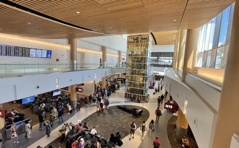Crowded airport concourse with departure screens, gate areas, and a multi‑story glass structure featuring yellow artwork.
