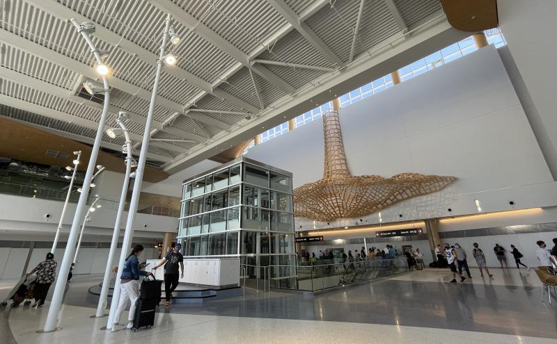 Spacious airport concourse featuring tall glass windows, a central glass elevator, and an overhead lattice-style wooden sculpture.