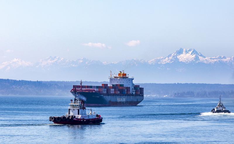 Hapag Lloyd container vessel in Elliott Bay guided by two tugs with snowy Olympic Mountains in the distance.