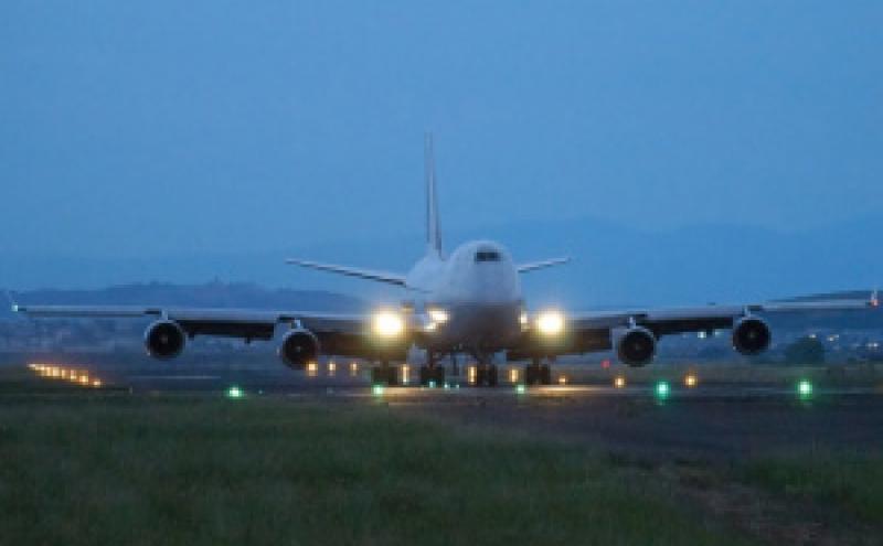 A large four‑engine airplane with its lights on is positioned on a runway at dusk, facing toward the viewer, with runway lights glowing and low mountains visible in the background.