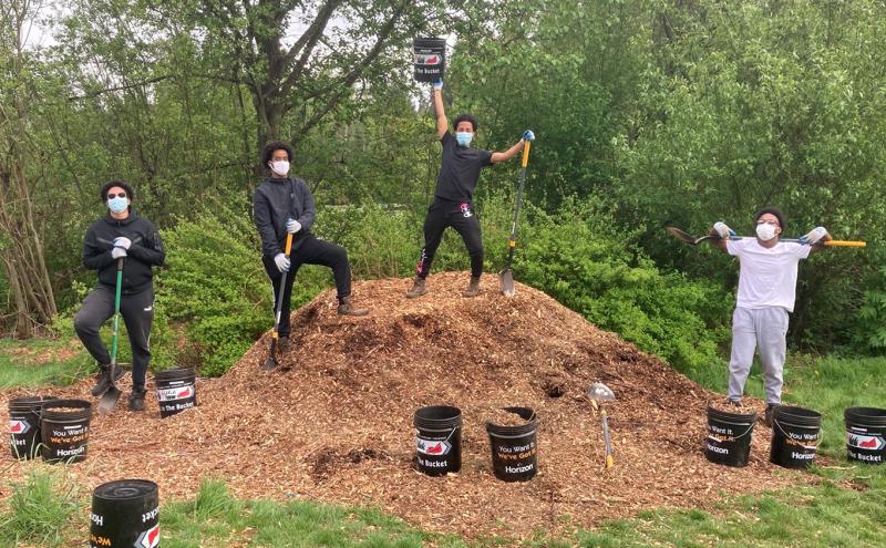 Four young men participants pose with shovels on and next to a pile of woodchips summer 2021