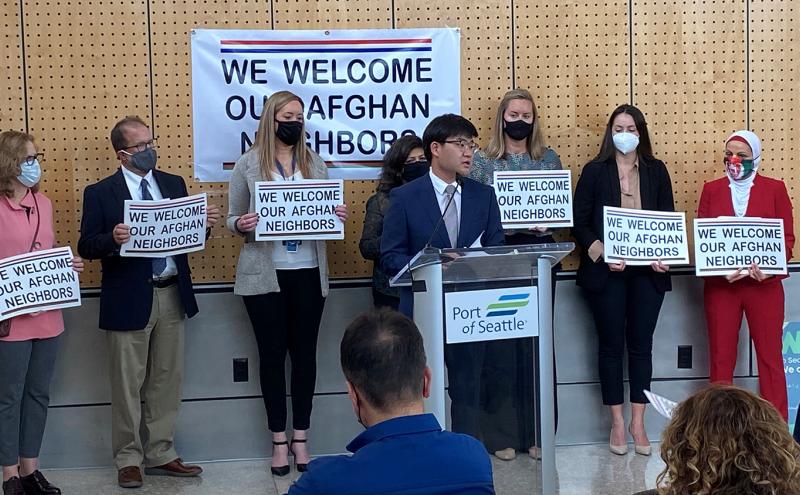 Commissioner Cho at a podium at SEA with a group lining the wall behind him holding supportive signs for Afghan refugees.