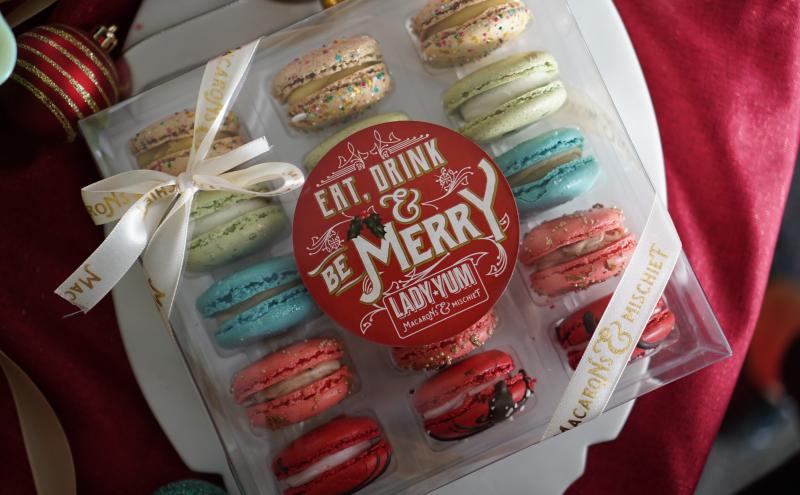 Box of assorted colorful macarons with a round red label reading ‘Eat, Drink & Be Merry,’ tied with a cream ribbon and displayed among holiday decorations.