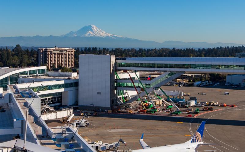 Airport terminal construction site with a skybridge and a snow‑covered mountain in the background.