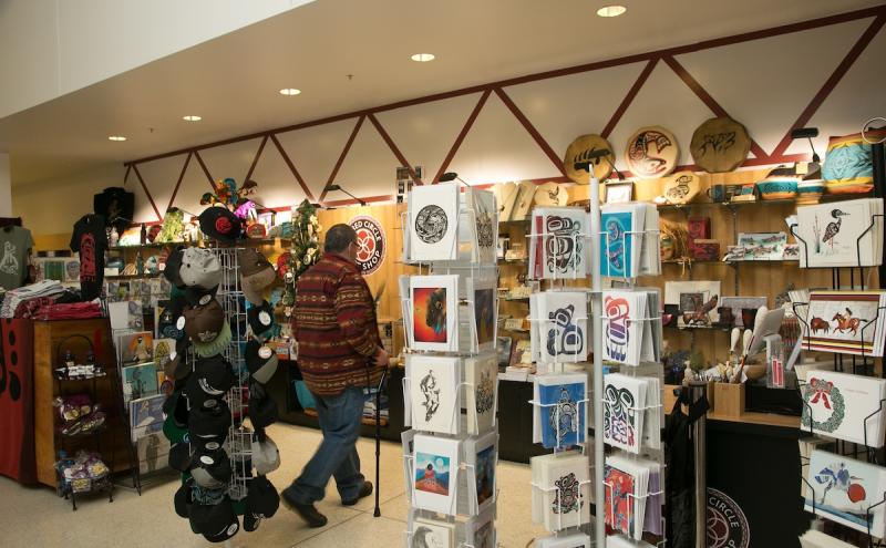 Sacred Circle gift shop with displays of Indigenous artwork, cards, hats, and souvenirs, with a shopper browsing the items.
