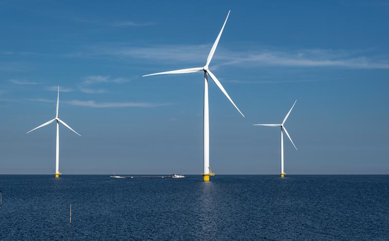 Three large offshore wind turbines stand in open water under a clear blue sky, with a vessel traveling across the horizon behind them.