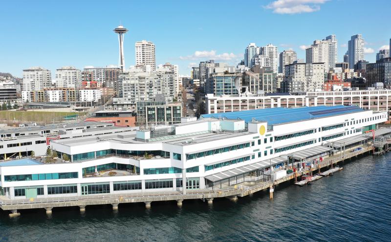 A large white waterfront building sits on a pier above the water, with the Seattle skyline and the Space Needle visible in the background under a clear blue sky.