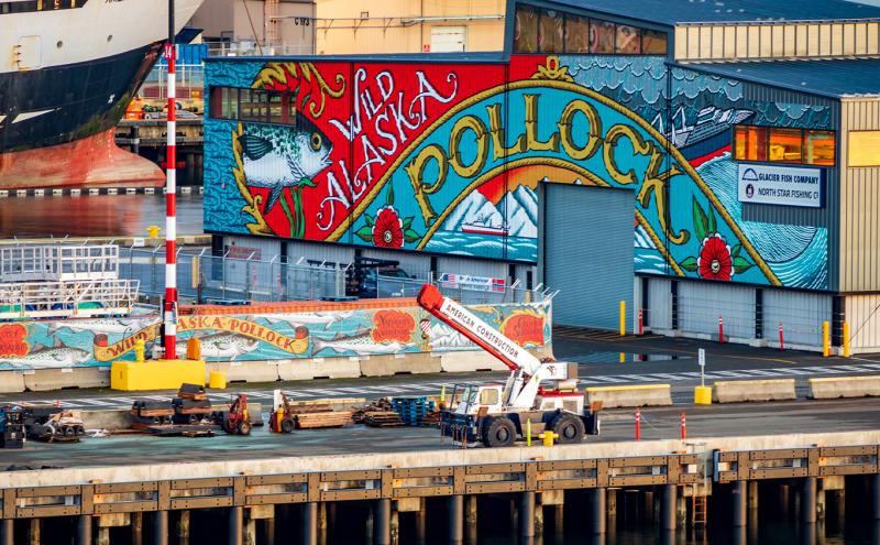 A colorful “Wild Alaska Pollock” mural on a waterfront building with a large ship and dock equipment nearby.