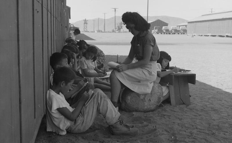At Manzanar Relocation Center California volunteer evacuee students and teachers sit on the shaded side of one of the barracks to work.