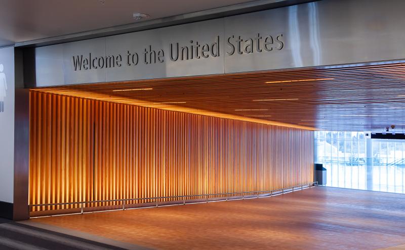 Interior walkway in an airport with a metallic sign reading ‘Welcome to the United States’ above the entrance. The hallway features vertical wooden slats illuminated with warm lighting along the right wall and ceiling. The floor transitions from a patterned surface into a brightly lit area with large windows visible in the background.