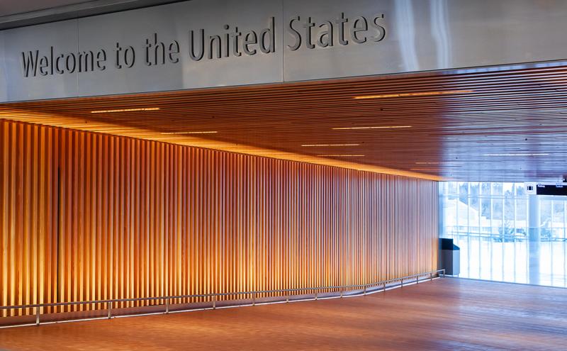 A modern indoor entry corridor with warm vertical wooden slats along the left wall, a wood-paneled ceiling with recessed lighting, and a sign above that reads ‘Welcome to the United States.’ Natural light enters through large windows at the far end of the hallway.