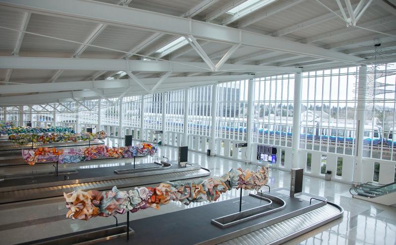 Bright, spacious airport baggage claim area with high white ceilings, large floor‑to‑ceiling windows, and multiple baggage carousels. Colorful abstract sculptural artwork is displayed above several carousels, stretching in long, ribbon‑like forms. A light‑rail train is visible outside through the windows, along with trees and distant buildings. Digital flight information screens and escalators are visible around the space.