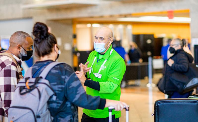 An airport staff member wearing a bright green shirt and a face mask speaks with travelers in a terminal area, gesturing with his hands as people wait in line with luggage.