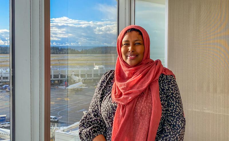 Ilays Aden smiling indoors with an airport runway and airplanes visible through the window behind them.