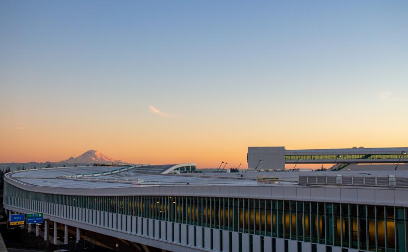 Sunset view of a modern airport terminal with long rows of windows and a curved roofline. The sky transitions from warm orange near the horizon to soft blue overhead. A snow‑covered mountain is visible in the distance behind the terminal. Highway signs and roadways run along the lower edge of the scene, with vehicles on the road.