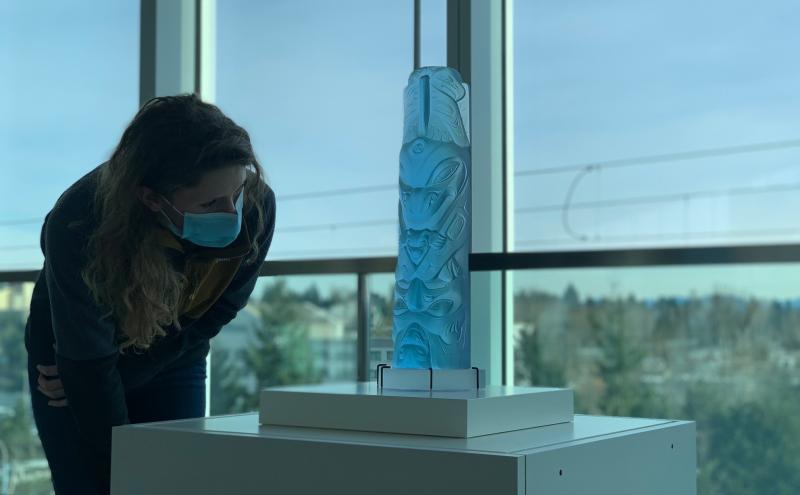 Person wearing a face mask leans forward to look closely at a light‑blue carved sculpture displayed on a white pedestal. The sculpture features intricate designs in a vertical column shape. The scene is inside a building with large floor‑to‑ceiling windows showing trees and distant buildings outside.