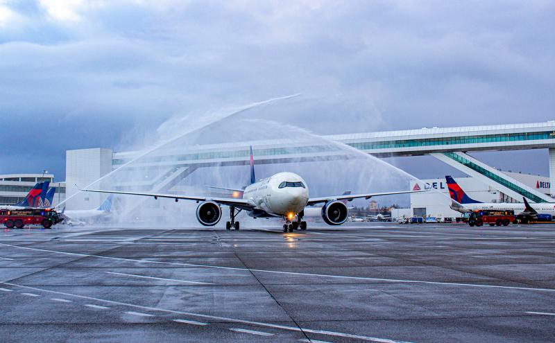 A commercial airplane on an airport tarmac passes beneath crossed streams of water sprayed by two fire trucks, creating a ceremonial water arch. The ground is wet, the sky is overcast, and airport buildings and additional aircraft are visible in the background.