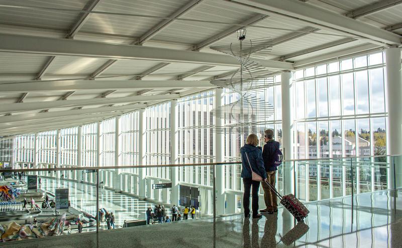 "Upper-level view inside a bright airport terminal with tall floor‑to‑ceiling windows and a high white ceiling supported by large beams. Two people stand near a glass railing, one pulling a rolling suitcase. Below them is the baggage claim area with multiple carousels and colorful sculptural artwork. A spiral wire sculpture hangs from the ceiling. Outside the windows, buildings and trees are visible under a partly cloudy sky.