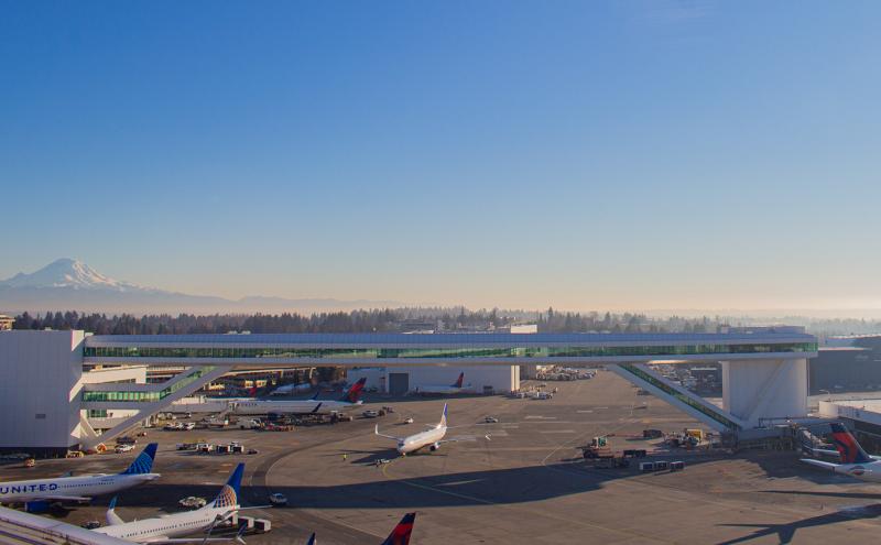 Wide view of an airport airfield with several parked commercial airplanes near terminal gates. A long elevated pedestrian walkway with glass walls stretches across the scene, connecting airport buildings. Ground service vehicles and workers are dispersed across the tarmac. In the distance, a tree-covered landscape and a large snow-covered mountain rise beneath a clear blue sky.